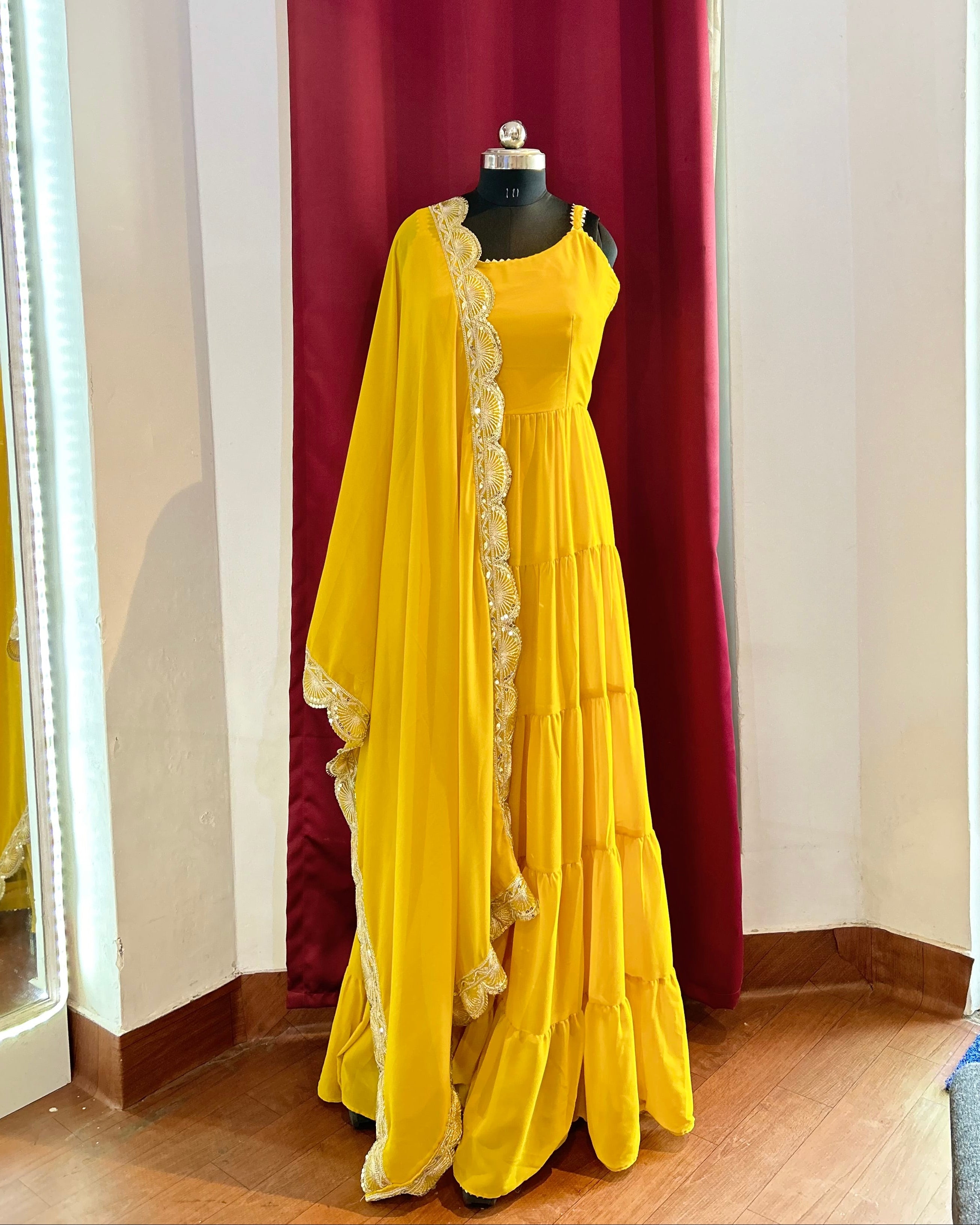 Yellow Anarkali with golden border on a mannequin against a red curtain.